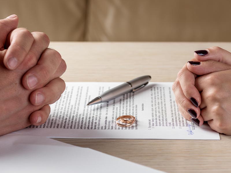 Man and woman's hands folded resting on divorce papers with wedding rings on paper